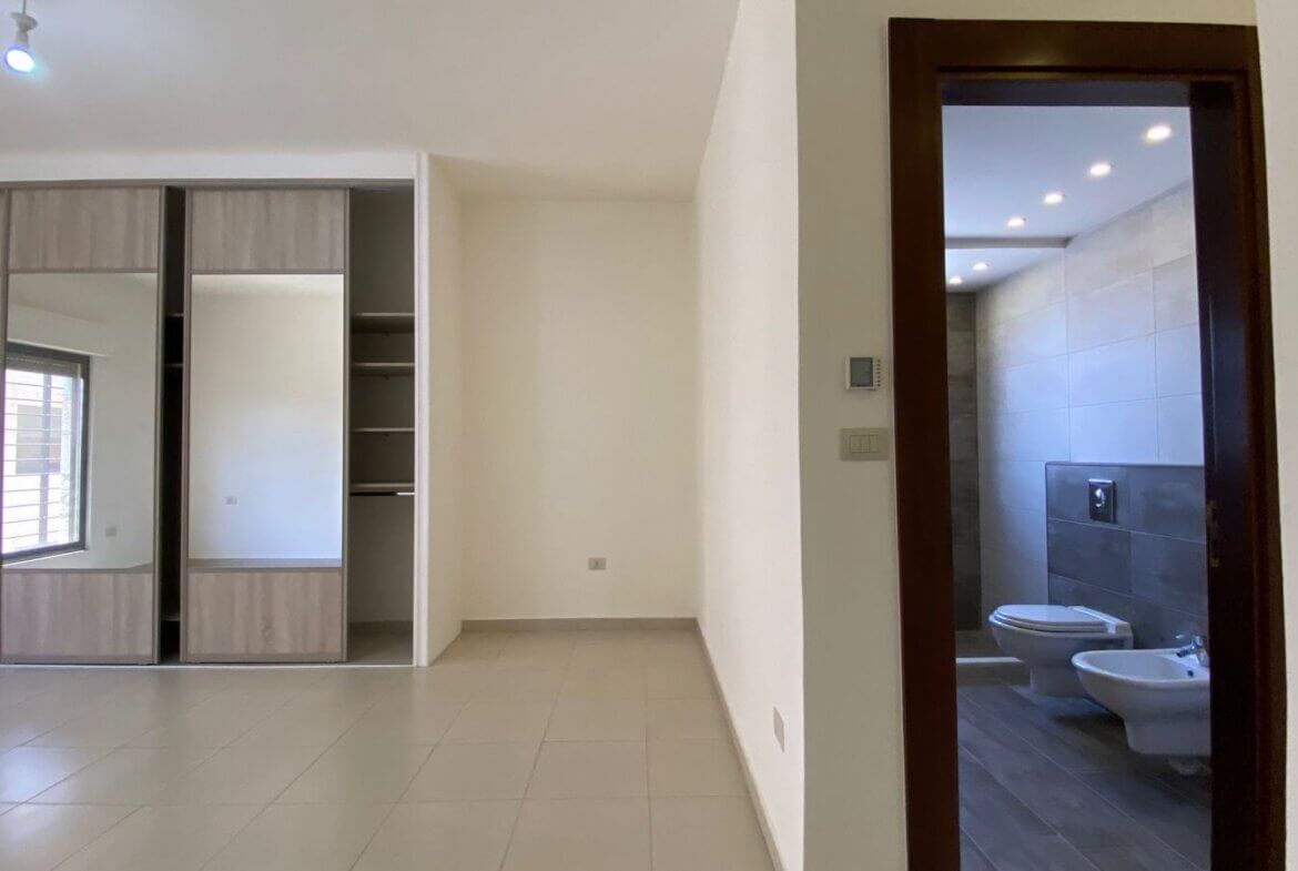 d02dcc92-ab16-4e2f-8253-2127b290f9bf Empty modern room with built-in wardrobes along the left wall and a dark wood doorway leading to a tiled bathroom with a toilet and bidet visible through the frame.