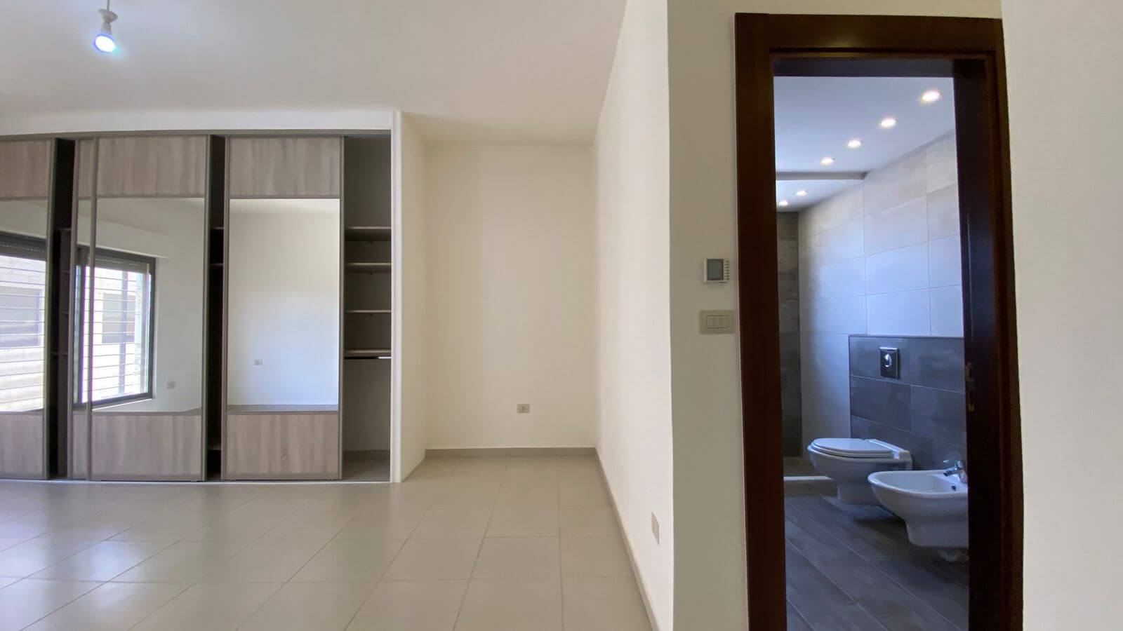 d02dcc92-ab16-4e2f-8253-2127b290f9bf Empty modern room with built-in wardrobes along the left wall and a dark wood doorway leading to a tiled bathroom with a toilet and bidet visible through the frame.