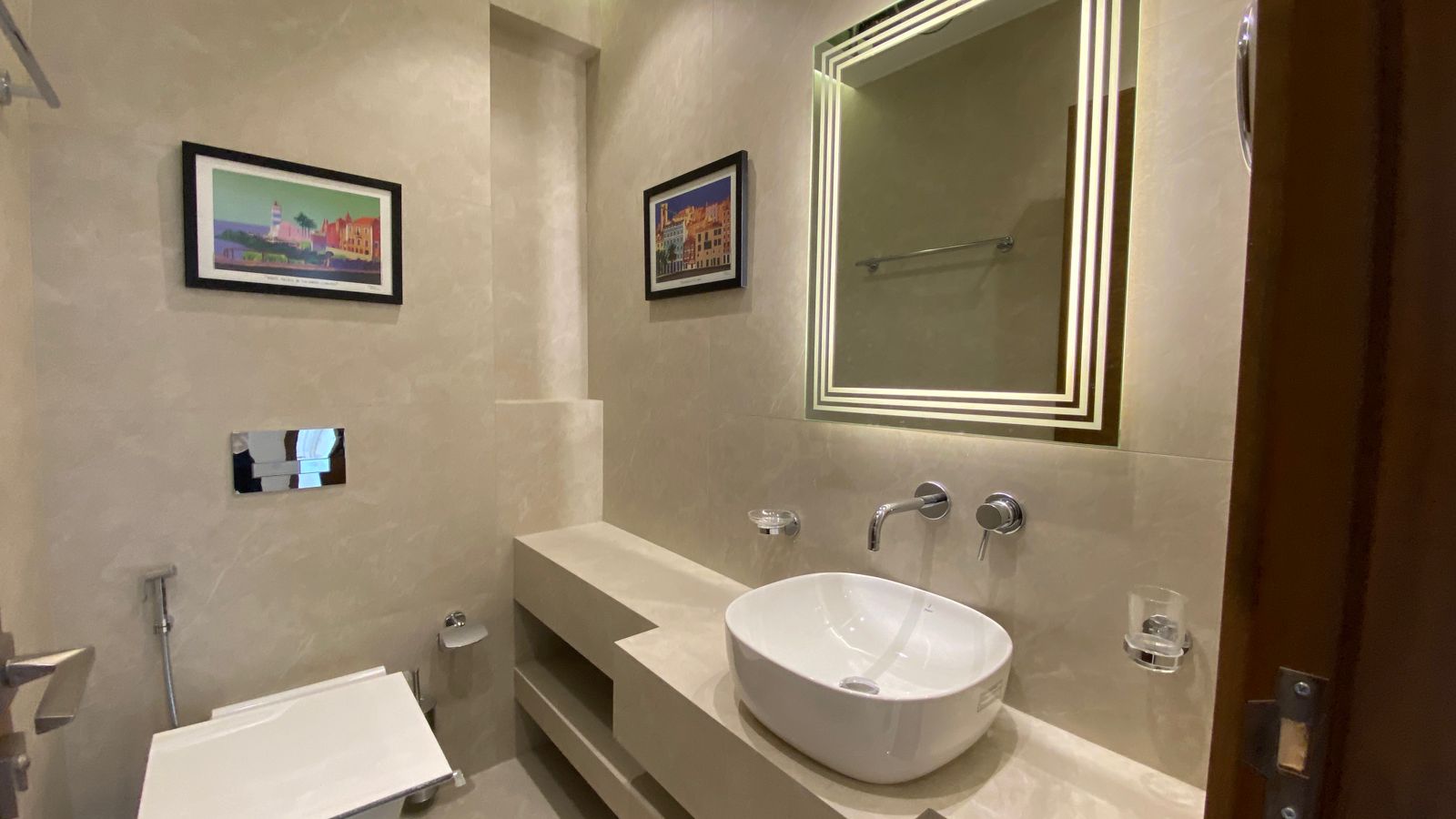 d1154eaa-4ff1-4c6c-a872-c2766243a7a3 Modern beige-tiled bathroom with a white vessel sink, wall-mounted faucet, and a backlit square-framed mirror with two framed pictures on the wall.