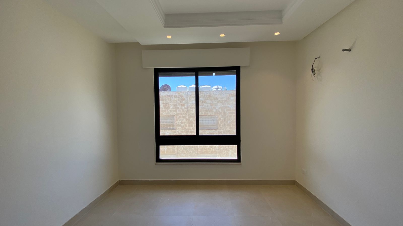 d2a77a44-ae33-4a0f-94fc-cdcc15961b8c Empty rectangular room with beige walls, tiled floor, and a large window overlooking a brick wall and rooftop water tanks outside.