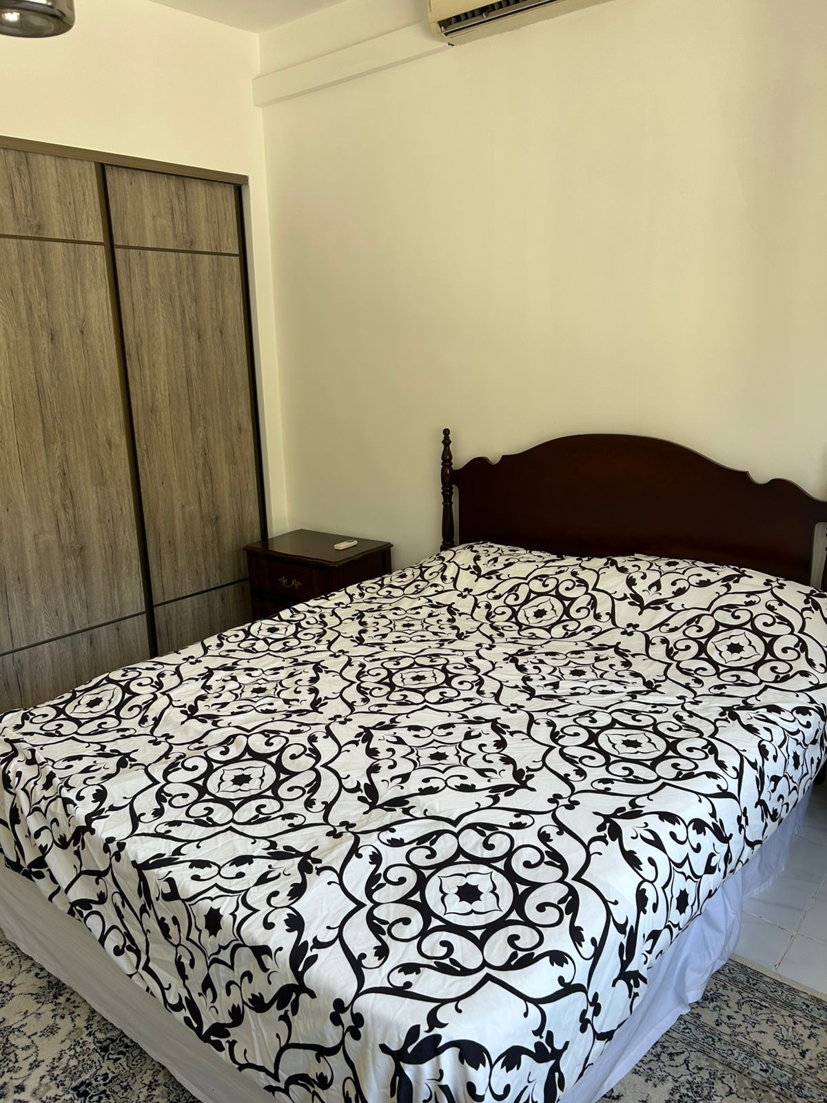 d3478f76-1201-4465-80cb-8f918562c333 A neatly made bed with a black and white patterned duvet, wooden headboard, and a small bedside table in a bright bedroom.