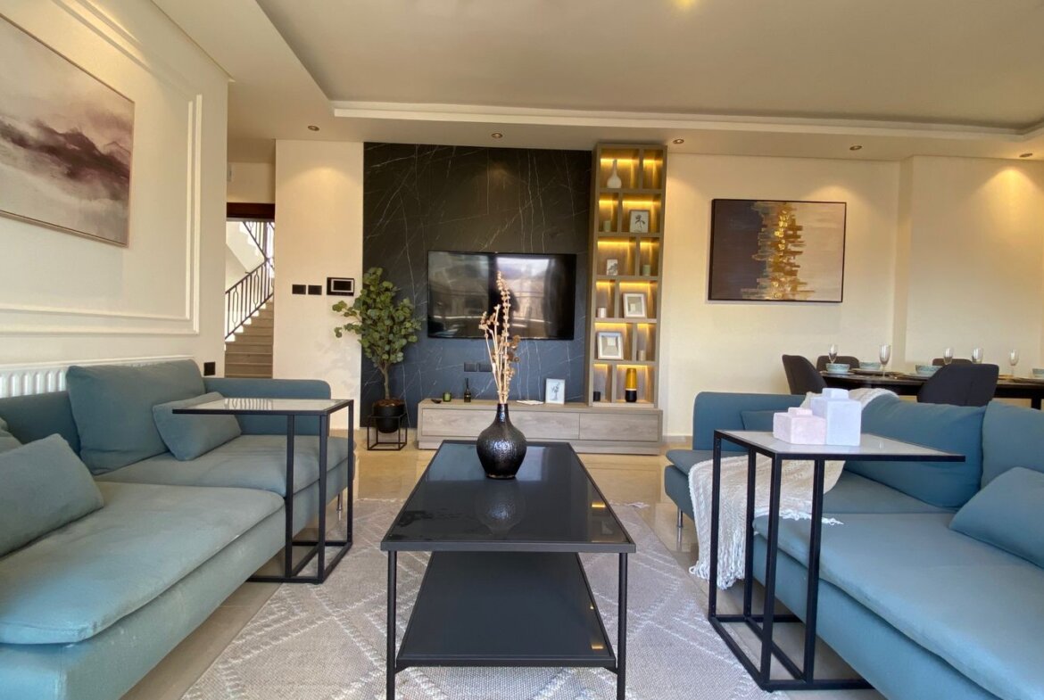 Modern living room with light blue sectionals, two glass-topped coffee tables, and a wall-mounted TV on a dark marble accent wall.