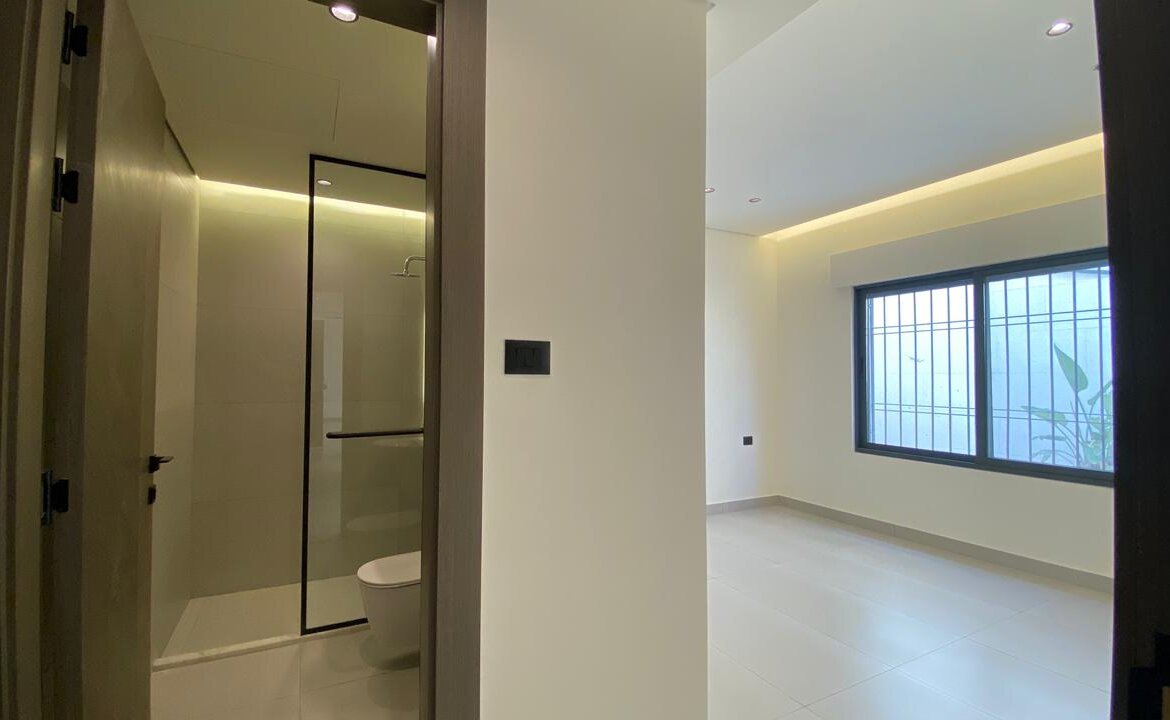 Modern, minimalist interior: a bright empty room with a large barred window, white walls and tiled floor; view into a bathroom with glass shower enclosure and toilet on the left.