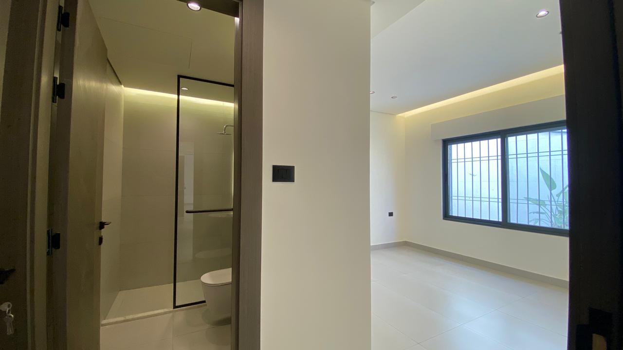 d35d48cb-cd58-49ca-8b03-9a8ddbab0889 Modern, minimalist interior: a bright empty room with a large barred window, white walls and tiled floor; view into a bathroom with glass shower enclosure and toilet on the left.