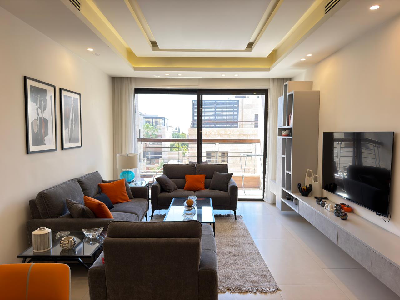 d4116559-01c5-4233-8aa8-0d04e6570872 Modern living room with gray sofas, orange cushions, a glass coffee table, and a wall-mounted TV facing large balcony doors.