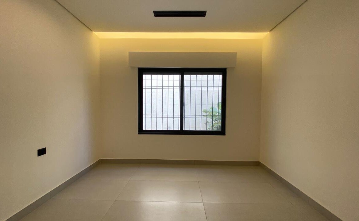 Empty rectangular room with beige walls, gray tiled floor, and a barred window on the far wall framed by warm ceiling LED lighting above. Outside the window is greenery visible through the bars.