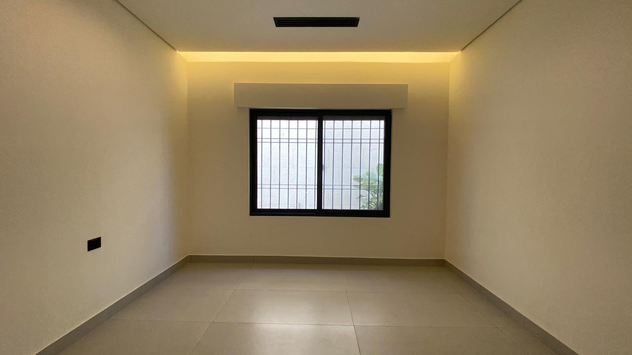 d4340b0d-46da-4e29-84af-d51d0241f493 Empty rectangular room with beige walls, gray tiled floor, and a barred window on the far wall framed by warm ceiling LED lighting above. Outside the window is greenery visible through the bars.