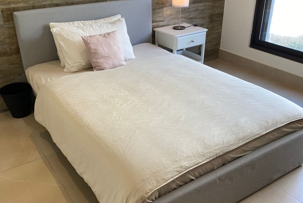 A modern bedroom with a low gray bed, white bedding, white pillows and a pink accent pillow against a wooden paneled wall, plus a white nightstand and lamp by the bed.