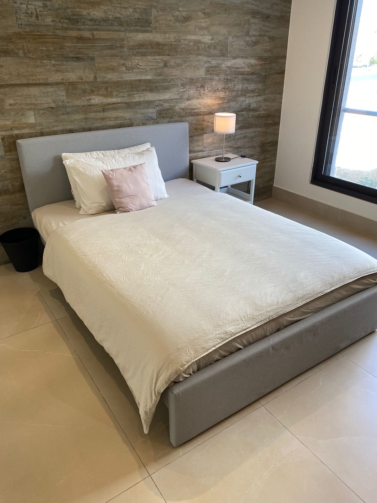 d4f2de0d-d08a-4a08-96b2-3abb750f608b A modern bedroom with a low gray bed, white bedding, white pillows and a pink accent pillow against a wooden paneled wall, plus a white nightstand and lamp by the bed.