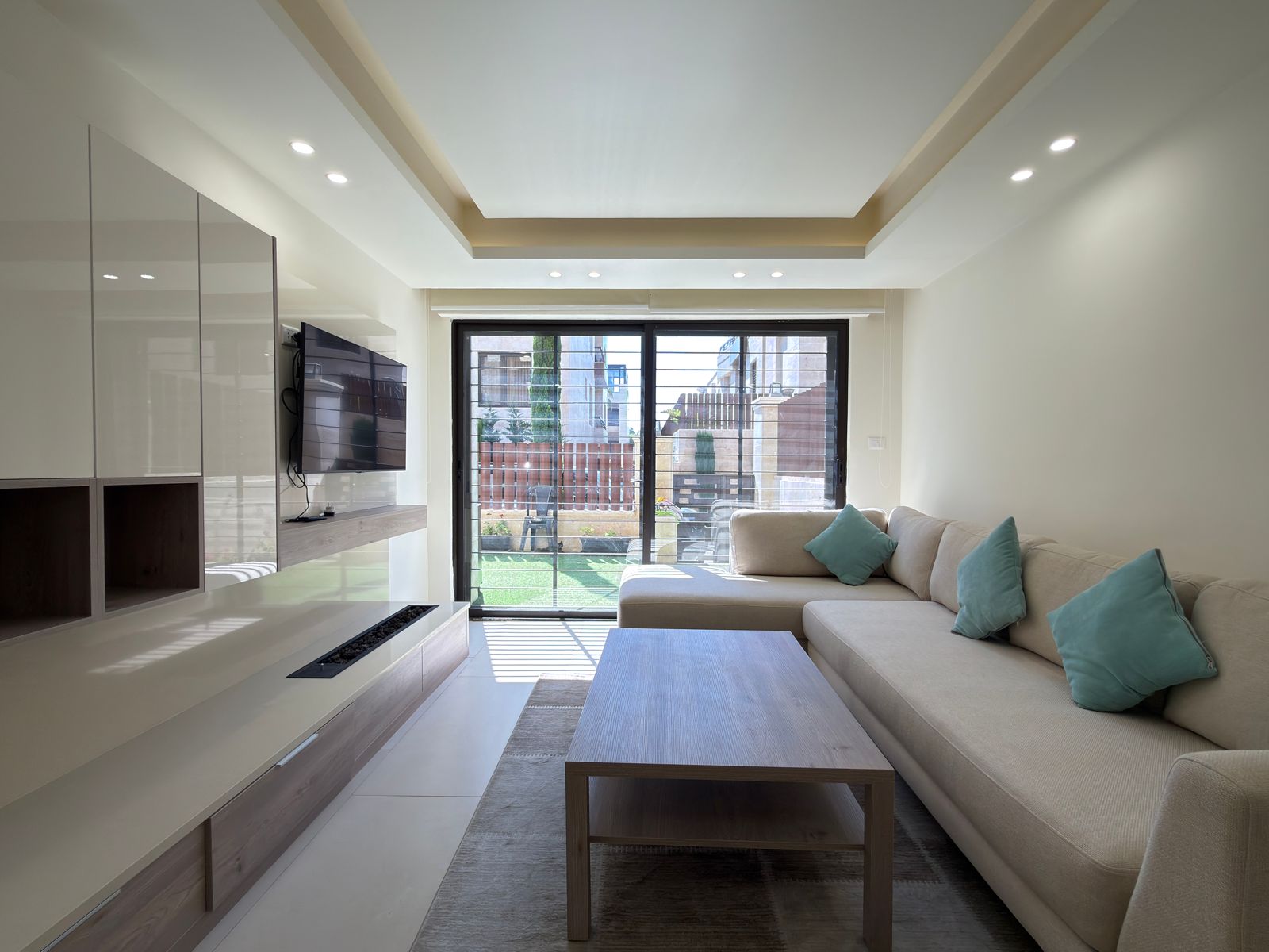 d5aeed70-004c-4e56-92f1-eaf665460211 Bright modern living room with a beige sectional sofa, turquoise cushions, and a wall-mounted TV opposite a sliding glass door to a fenced backyard outside.