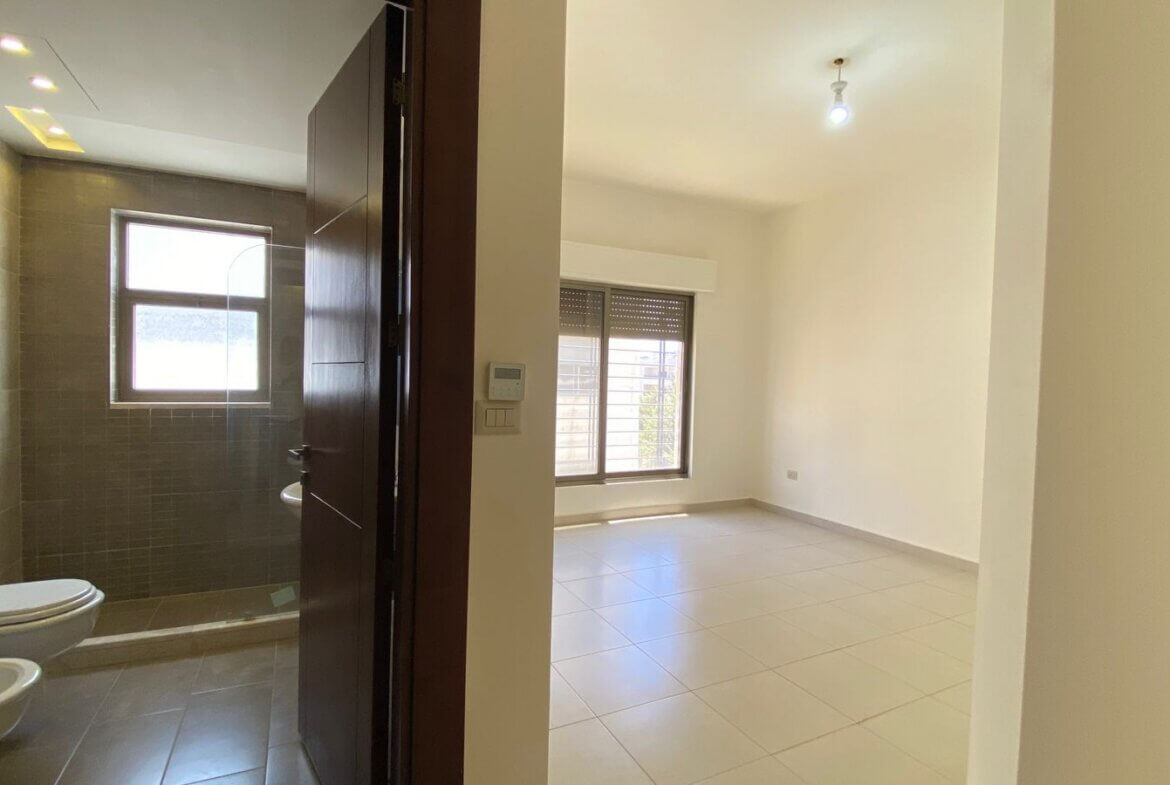 d6359b07-634f-48a7-9a88-288ade4f0160 A doorway opens into a bathroom on the left with a toilet and bidet; a window above tiled walls, and an adjacent bright empty living room with a large window and tiled floor on the right.