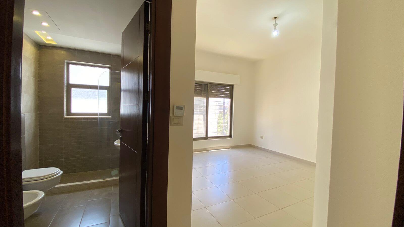 d6359b07-634f-48a7-9a88-288ade4f0160 A doorway opens into a bathroom on the left with a toilet and bidet; a window above tiled walls, and an adjacent bright empty living room with a large window and tiled floor on the right.