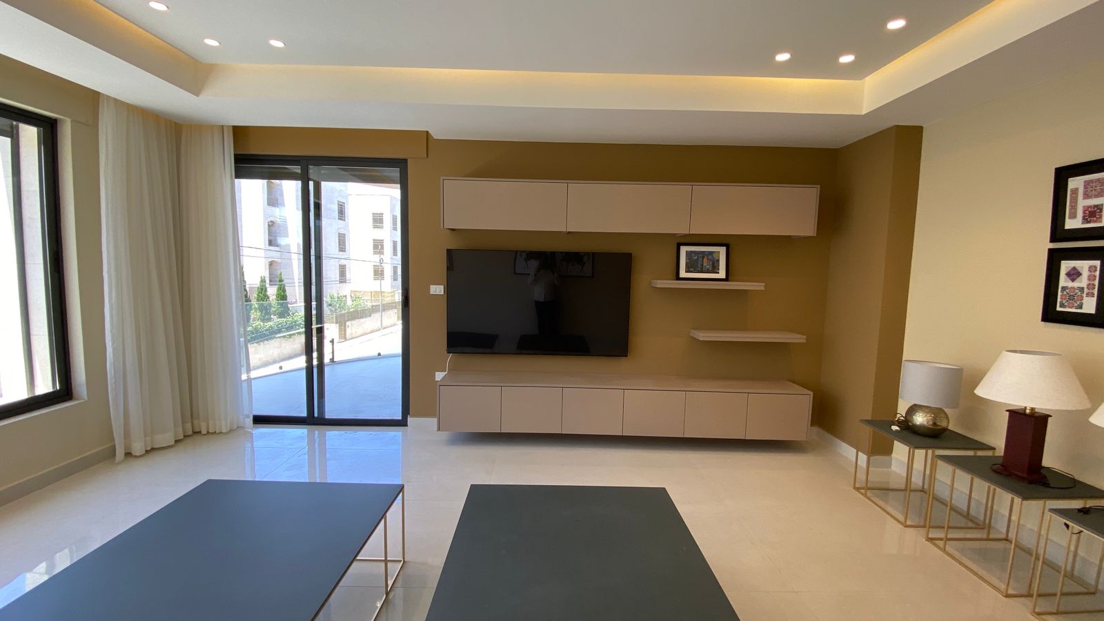 d97a78b9-5ddd-4730-9ffb-d18cbb2fdc96 Modern living room with a wall-mounted flat-screen TV, beige storage cabinets and shelves on a brown accent wall, a sliding glass door to a balcony, white curtains, and two dark gray coffee tables.