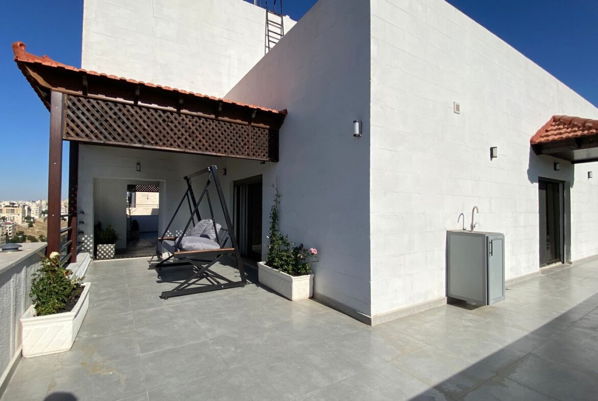 Rooftop terrace with a wooden lattice pergola, a swinging chair, and potted plants against a white building under a clear blue sky.