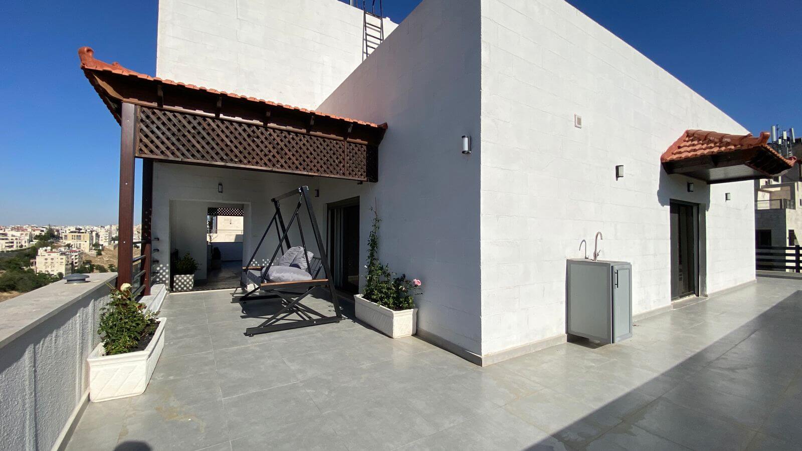 Rooftop terrace with a wooden lattice pergola, a swinging chair, and potted plants against a white building under a clear blue sky.