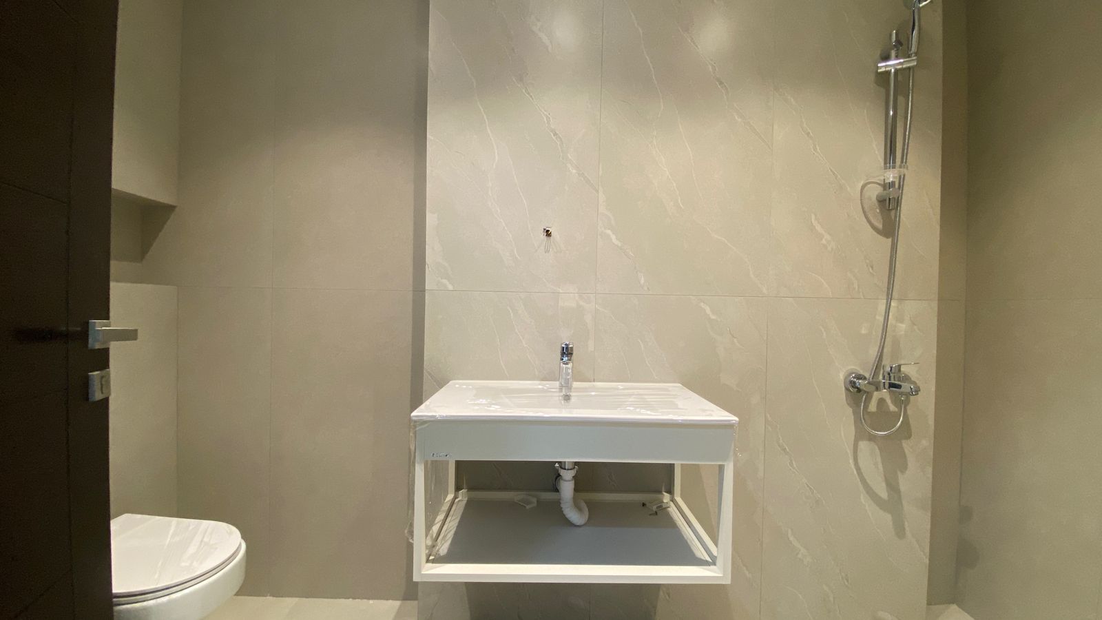 dab0c941-4671-41f9-93c0-9660bd40402e Modern bathroom with wall-mounted sink, exposed plumbing, and a shower attachment on the right; toilet visible on the left.