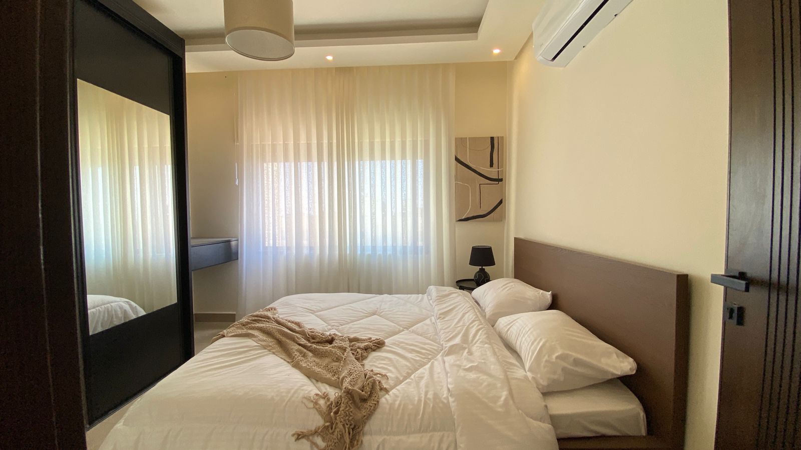 db99ac17-7530-46bd-bb13-5fabc814668b A modern bedroom with a low wooden headboard, white bedding, and a beige throw on a neatly made bed; a black bedside lamp on the right and sheer curtains over a window in the back.
