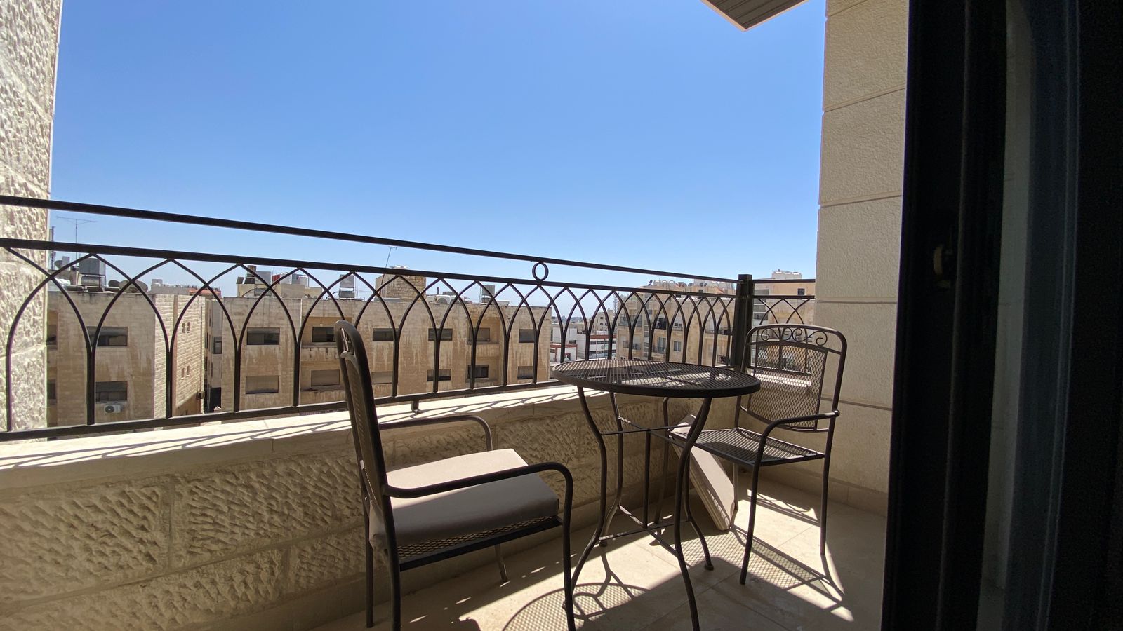 db9cbcdf-7160-43bc-9c40-9be14d23976d Balcony with a metal table and two chairs on a sunlit terrace overlooking beige city buildings under a clear blue sky.