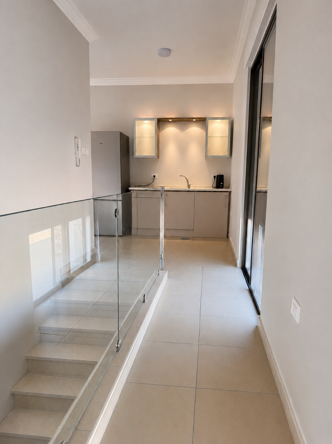dcbe9b7d-6d0e-4137-b22b-2b5de950447e Compact modern kitchenette at end of a bright hallway: grey cabinets, stainless fridge, sink, and glass-front wall cabinets with under-cabinet lighting.