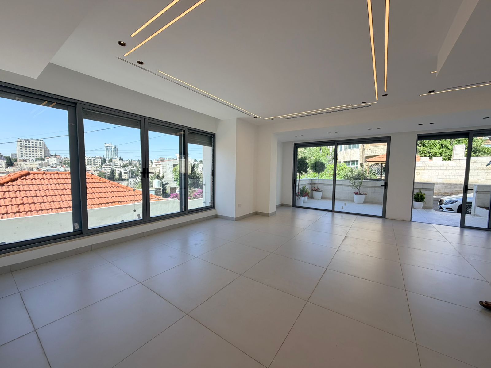 dcffa50d-9c25-4bb4-aee4-1720f9e3ed1b Bright empty living room with large floor-to-ceiling windows and sliding glass doors to a balcony, city view outside.