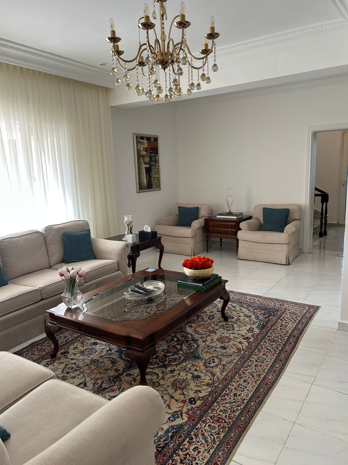 dd2f5c97-4749-4889-ab29-e55d8c4d46c0 Luxurious living room with beige sofas, teal cushions, and a glass-topped wooden coffee table on a patterned rug under a crystal chandelier