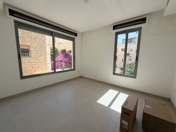 Empty sunlit room with large sliding windows showing stone buildings and pink bougainvillea outside; a few cardboard boxes on the tiled floor.