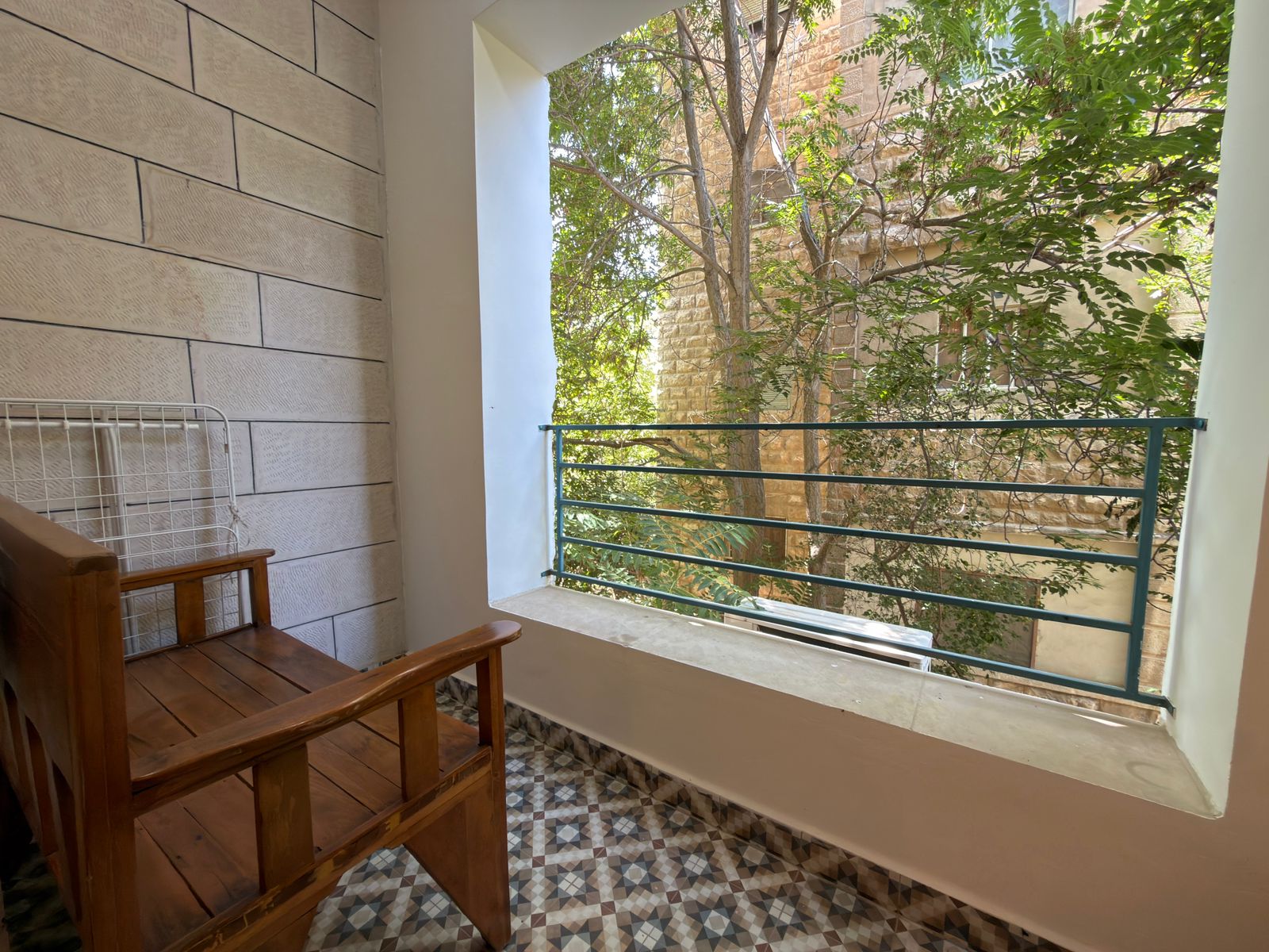ded39dbd-f253-4c1c-9608-56c29d66cdf2 Wooden bench on a patterned tile balcony with a metal railing and leafy trees outside the wall.