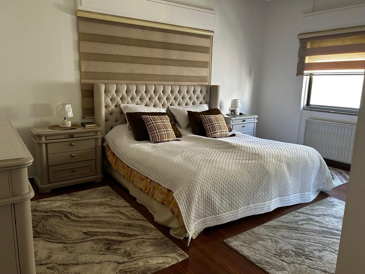 df408ee6-08c0-4db1-9e35-8a446407bcbf Queen-sized bed with a tufted beige headboard, white quilted cover, and brown patterned throw pillows flanked by matching nightstands with lamps.