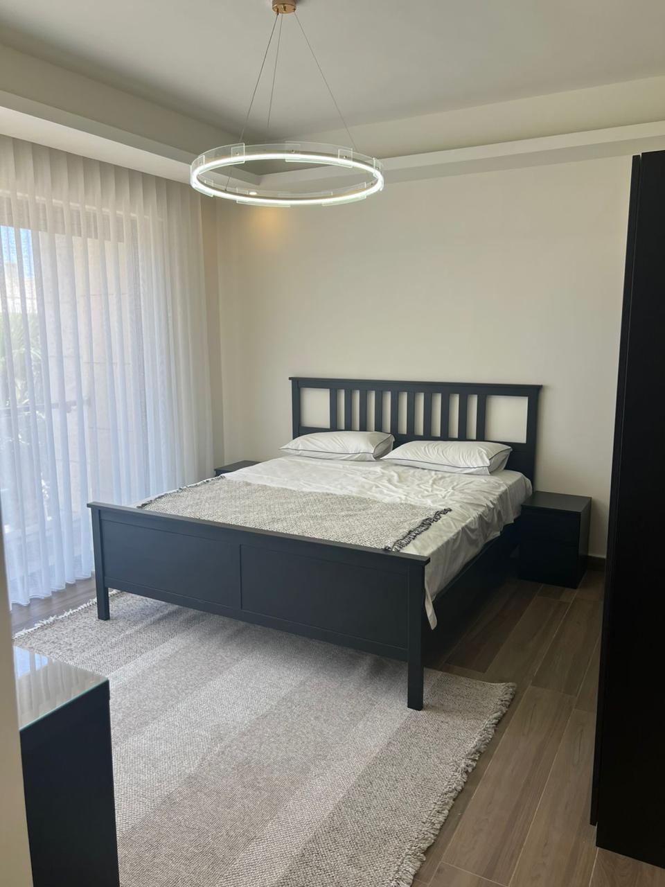 df6516db-812f-46b8-836c-7db457c88884 Black wood queen bed with white sheets and pillows, circular halo ceiling light, two black nightstands, and a beige rug in a minimalist bedroom