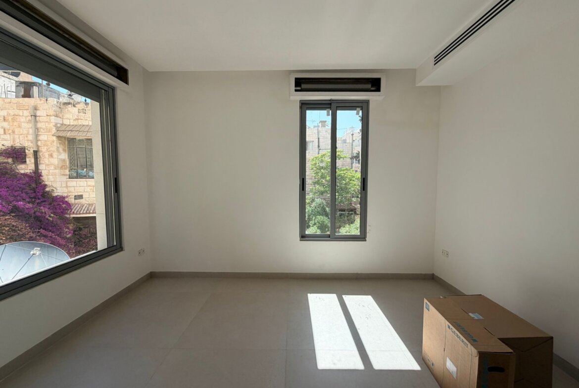 Empty rectangular room with large side window and a smaller central window, sunlight casting shadows; a cardboard box sits on the tiled floor nearby.