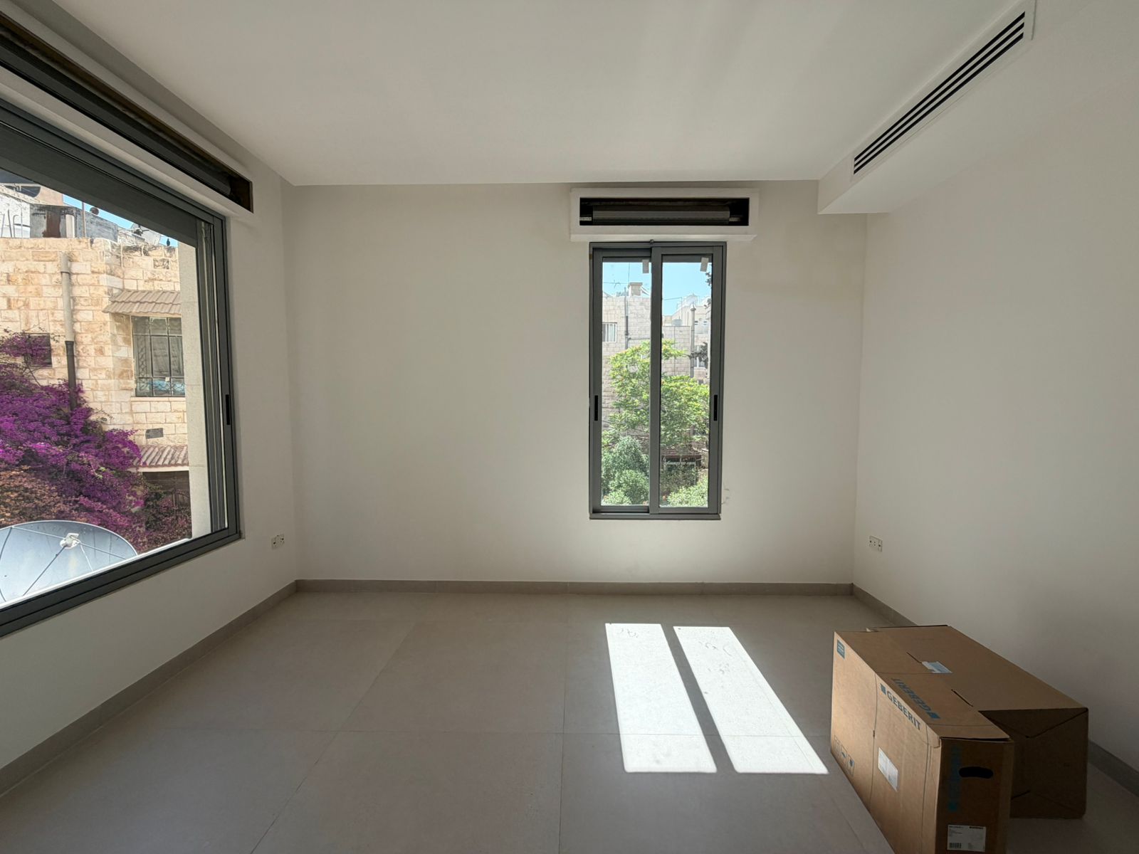 dfbfacee-7cc1-441f-a4b3-3aa2170f1ddb Empty rectangular room with large side window and a smaller central window, sunlight casting shadows; a cardboard box sits on the tiled floor nearby.