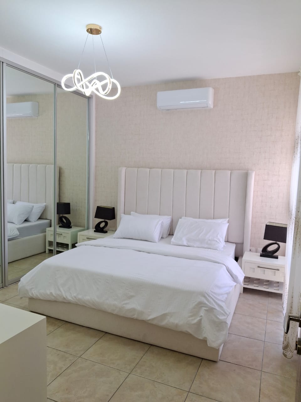 e08e0db7-8877-420f-96c5-55bfcf4556bb Bright modern bedroom with a white upholstered double bed, matching nightstands, black lamps, mirrored wardrobe, and pendant chandelier to the left.