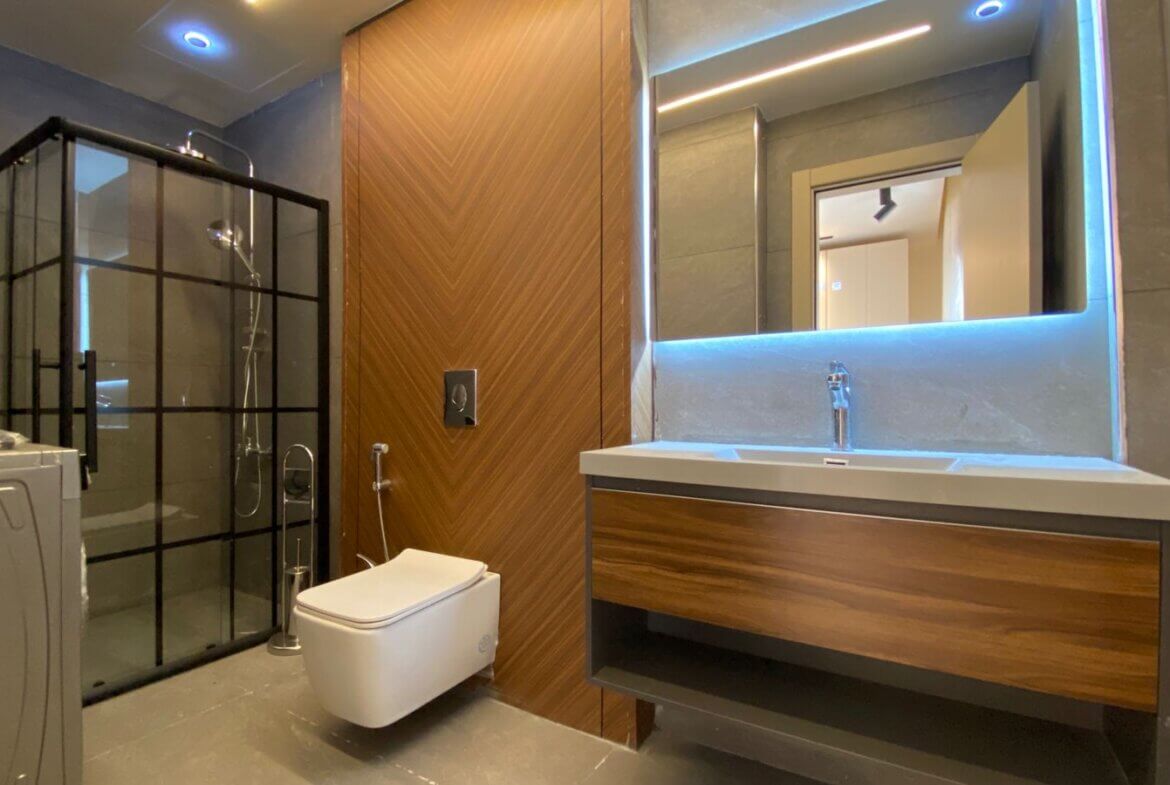 Modern bathroom with a glass-framed shower on the left, a wall-mounted toilet, and a wooden vanity with a backlit mirror above the sink.