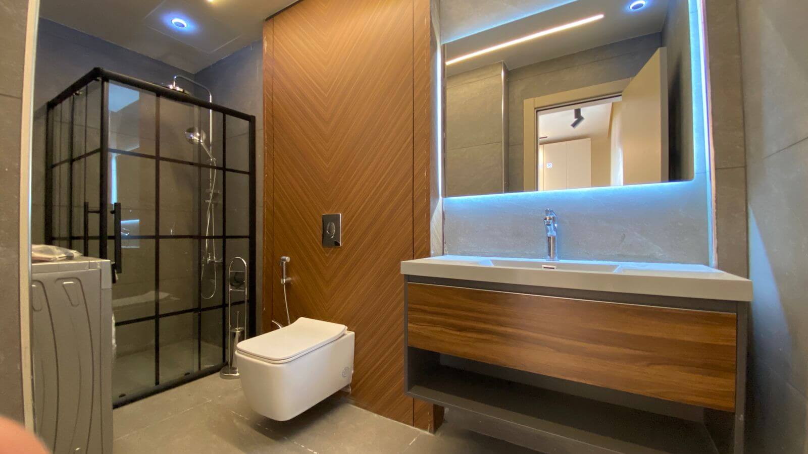 Modern bathroom with a glass-framed shower on the left, a wall-mounted toilet, and a wooden vanity with a backlit mirror above the sink.