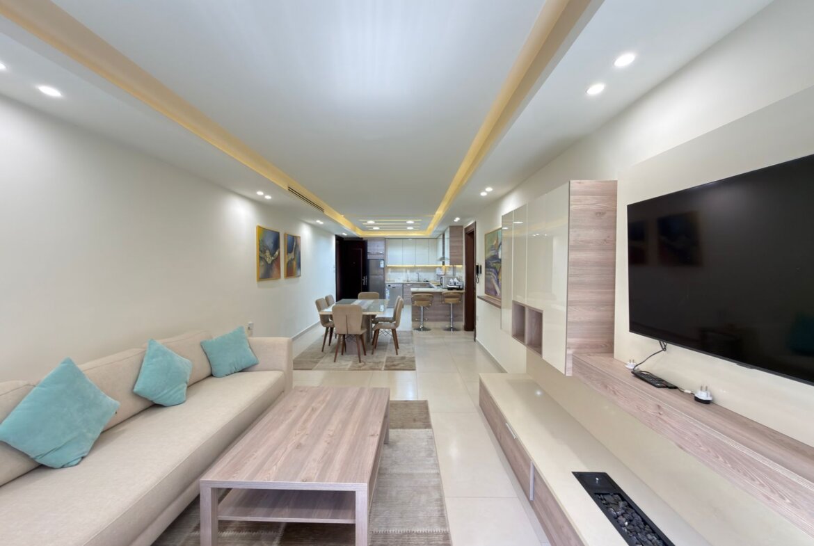 Open-concept living room with a beige sofa and turquoise cushions, a wooden coffee table, and a large TV; dining area and kitchen visible in the background.
