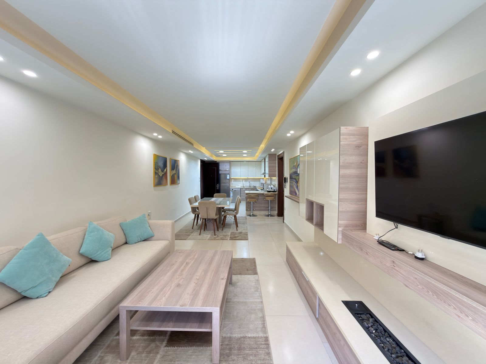 e4166084-956a-4439-9aab-0f34bed7a35f Open-concept living room with a beige sofa and turquoise cushions, a wooden coffee table, and a large TV; dining area and kitchen visible in the background.