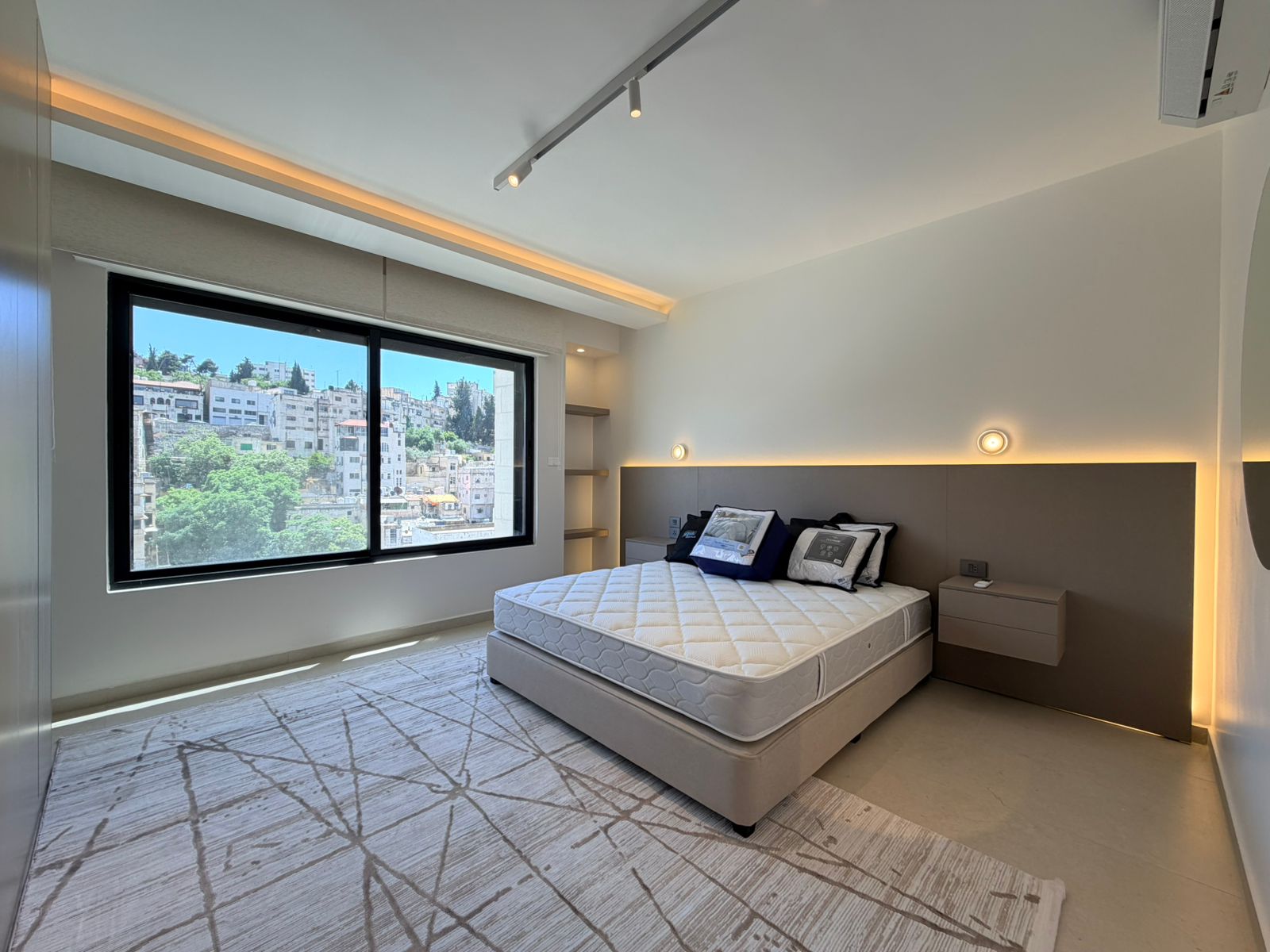 e42723dd-57ad-4ccb-b780-0eab49ac8b2b Modern bedroom with a king bed, gray headboard, two floating nightstands, warm backlighting, and a large window overlooking a hillside cityscape.