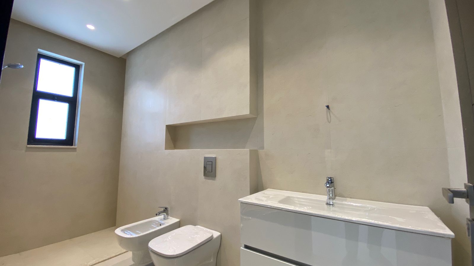 e4d5e5bb-e294-4788-8507-e1067afa7969 Modern beige bathroom with a wall-mounted sink, toilet and bidet, and built-in shelves by a tall wall window.