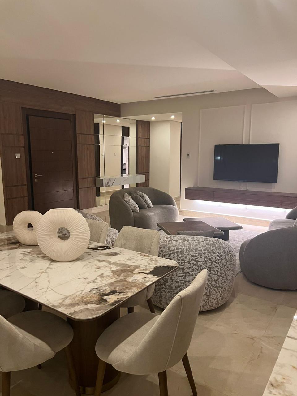 e68a9a23-331f-40d5-957b-b562c921deec Open-plan living and dining area with a marble dining table, beige chairs, gray curved sofas, and a wall-mounted TV.