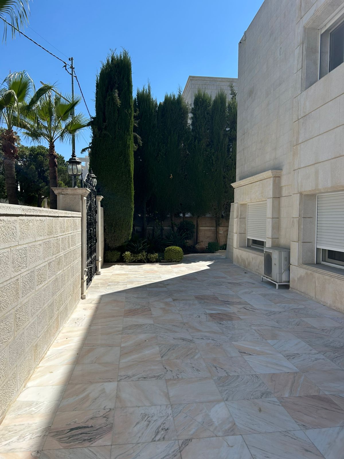 e745ad89-9d8e-48eb-837a-64061bbb398f Sunny marble courtyard beside a beige stone building, with a wrought-iron gate and tall evergreen trees along the wall.