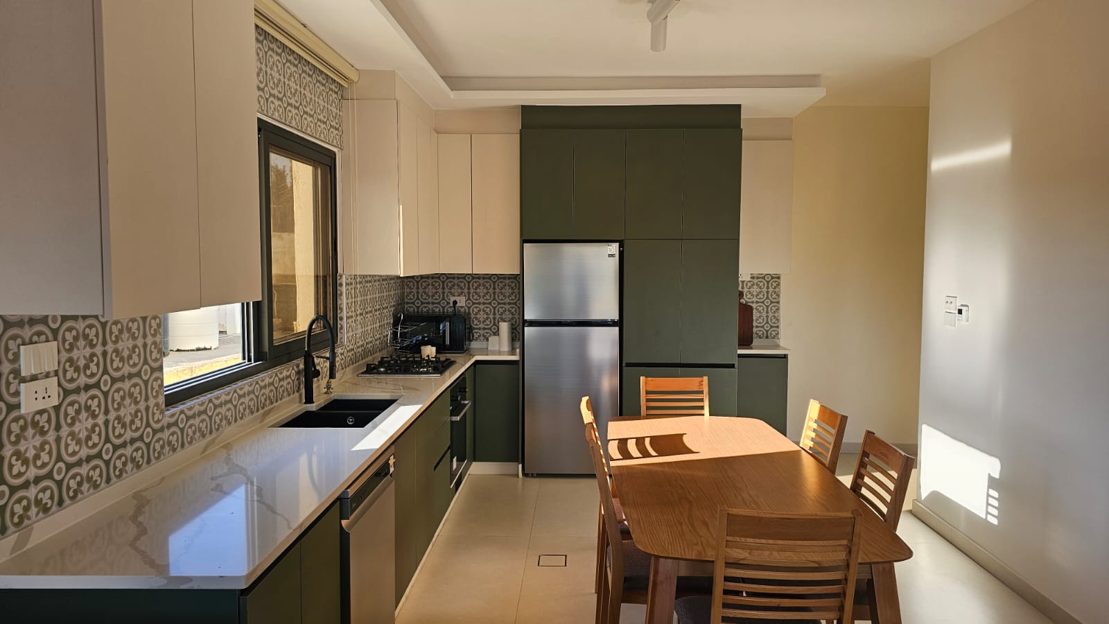 e7b0e97f-0a40-45ef-b8fd-469a50206e85 Modern kitchen with green lower cabinets, white countertops, stainless fridge, and a wooden dining table nearby.