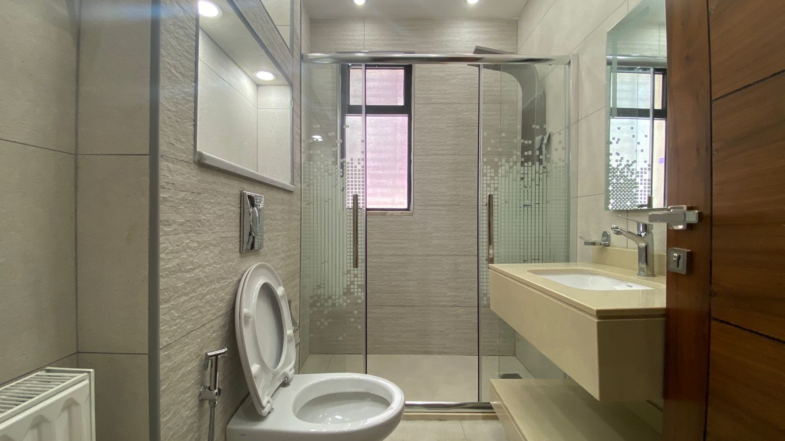 e8a4f4aa-61b5-480e-90ec-28fa3ae5cdb9 Modern bathroom with a glass-framed shower, toilet, and beige wall-mounted sink in a neutral-toned space.