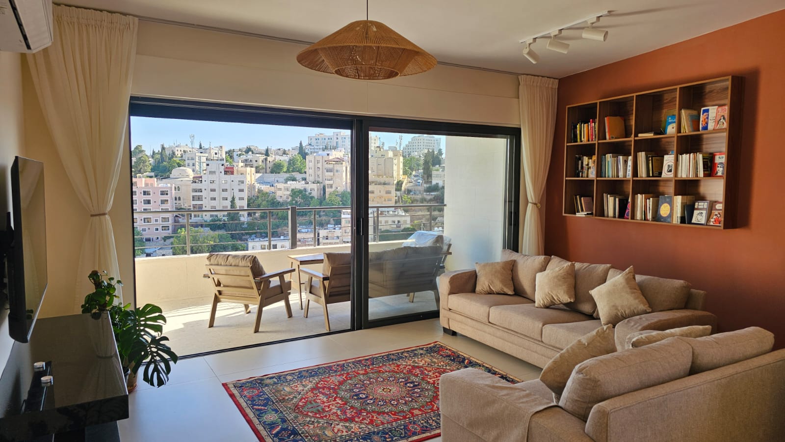 e8f8711d-26e5-4c4d-a71c-8441a27c79a5 Living room with beige sectional sofa, bookshelf on an orange wall, and a colorful rug facing a sliding glass door to a balcony with city view. a wicker pendant light hangs from the ceiling.