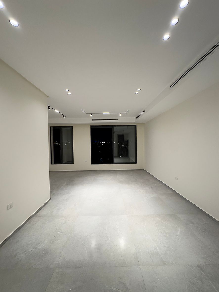 e946b442-a3a8-4c40-a798-18a2092854c9 Empty modern living room at night with large windows, white walls, gray tiled floor, and recessed ceiling lights.
