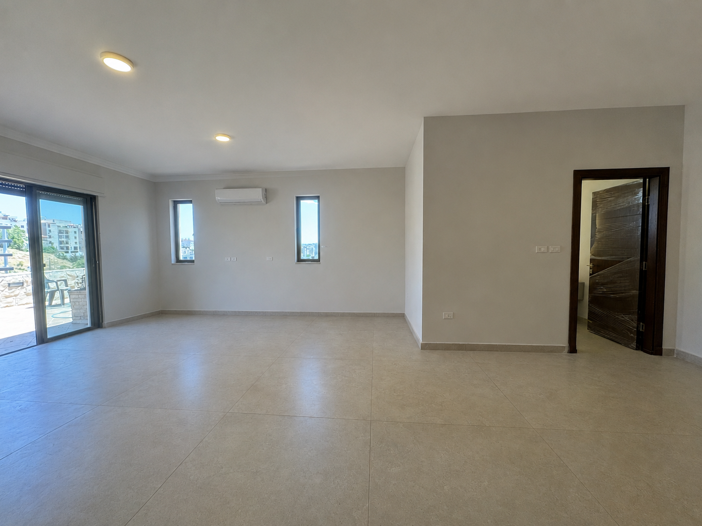 e9ece4a0-b422-4123-8ab1-a733a8690a16 Empty living room with beige tiled floor, large sliding glass door to a balcony on the left, and two narrow vertical windows with a wall-mounted air conditioner between them.