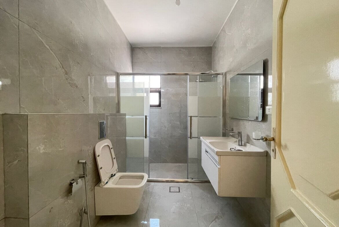 ea005ca9-6c45-4dcf-aa74-102dc3e0fa38 Modern grey-tiled bathroom with a wall-mounted toilet, frosted glass shower enclosure, and white vanity with sink against the right wall; small window visible in shower area.