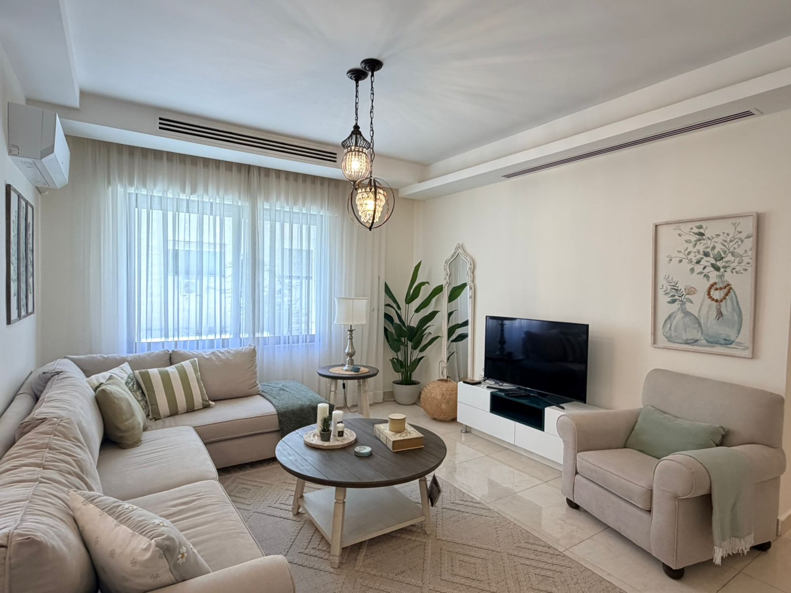 eb4edd6b-d5d8-4add-b4b6-e0f20f3430b7 Bright living room with a beige sectional, sheer white curtains, and a TV on a white media console.