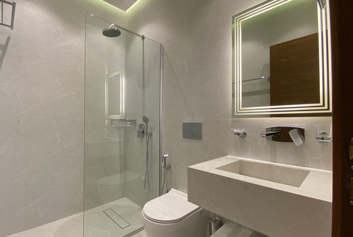 Modern bathroom with glass-enclosed shower, wall-mounted toilet, and a square backlit mirror above a concrete sink.
