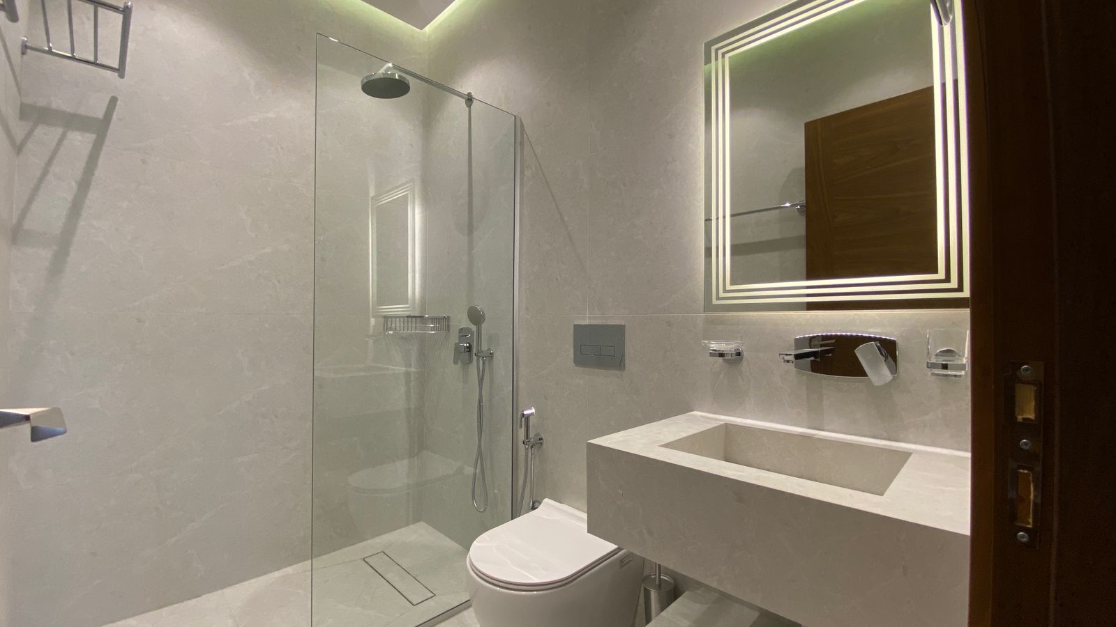 eb67a808-35e2-4859-84ec-22ca39fe60e0 Modern bathroom with glass-enclosed shower, wall-mounted toilet, and a square backlit mirror above a concrete sink.