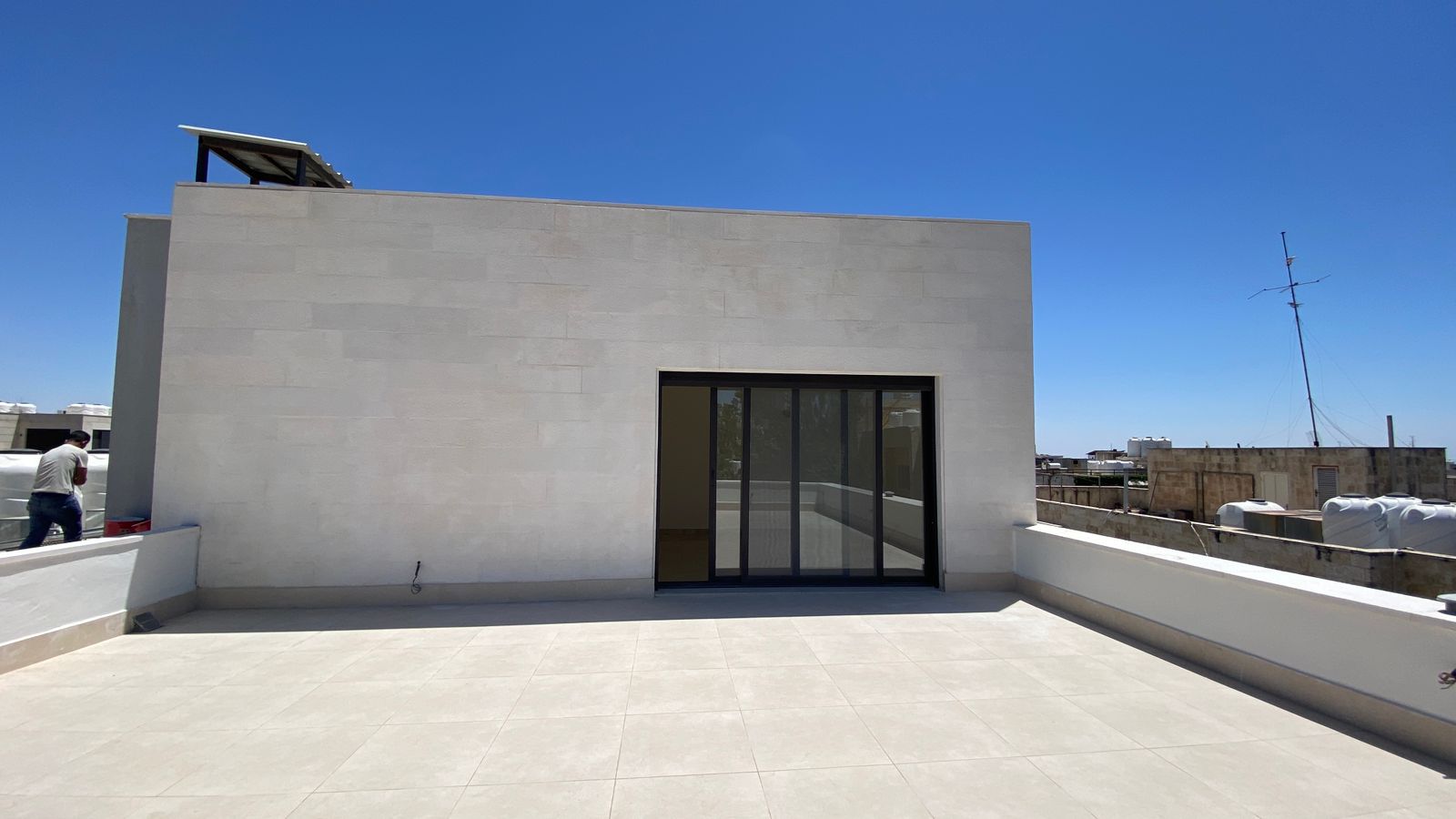 eb8503c3-5d16-45b4-b531-4f59559ff98d Rooftop terrace with a large gray wall and sliding glass door, clear blue sky, and a man near the left edge.